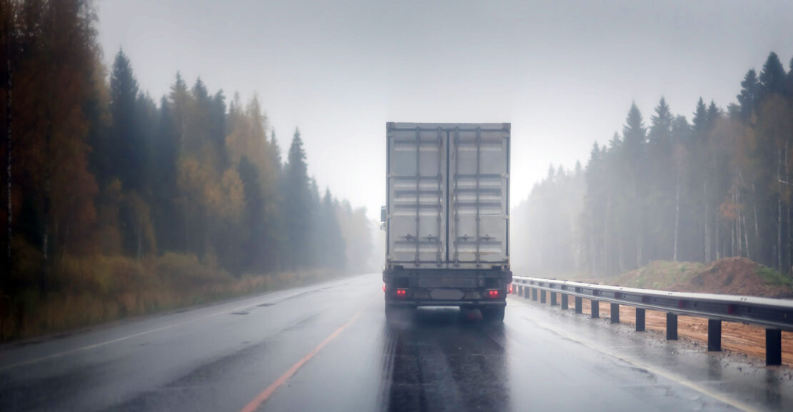 Big rig semi truck with refrigerator unit on reefer trailer transports commercial industrial cargo on a multi-lines highway with wet shiny coating and rain dust in rainy weather.
