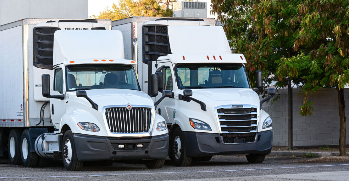 Two white semi-trucks parked at large warehouse dock