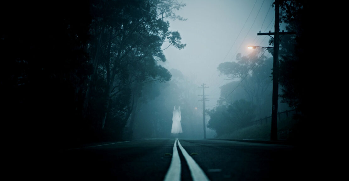 A dark, foggy night, with a female ghost standing in the middle of the road off in the distance