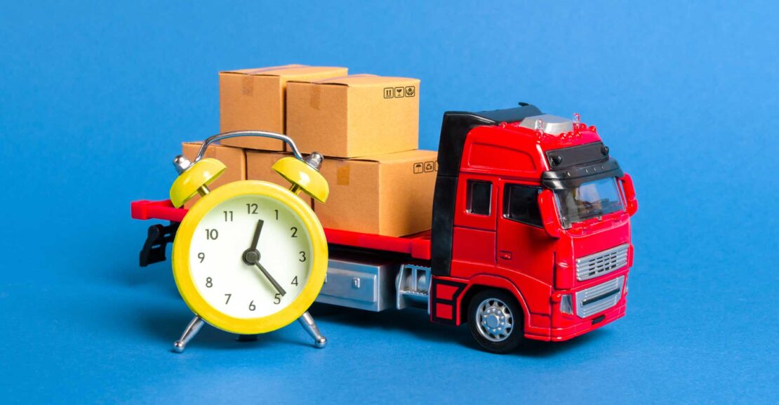 A toy red flatbed truck carrying boxes in front of an alarm clock