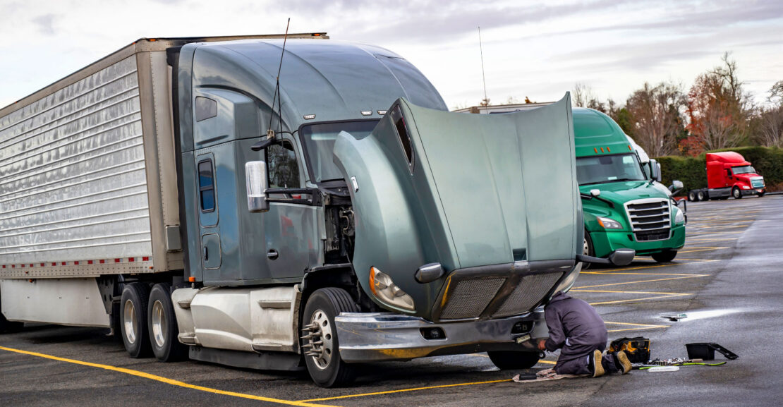 Truck driver repairing broken gray big rig semi truck with open hood and refrigerator semi trailer directly at the truck stop parking lot